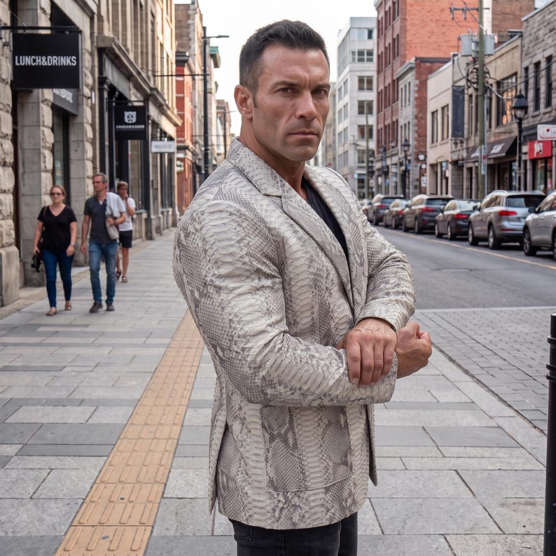 May include: A light grey and white blazer featuring a detailed snakeskin pattern throughout. The garment has a notched lapel, long sleeves, and two front patch pockets. The blazer is worn over a plain black shirt and paired with dark trousers.