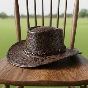 May include: A brown leather hat with a snakeskin pattern rests on a wooden chair. The hat has a wide brim and a braided band around the crown. The hat features small metal eyelets and studs. The chair is made of wood.