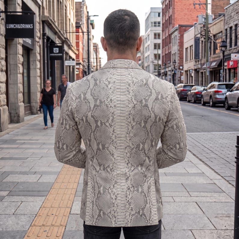 May include: A blazer featuring a light beige and grey snakeskin pattern. The garment has a structured collar, long sleeves, and a tailored fit, shown from the back. The material displays a detailed, repeating scale texture across the entire surface.