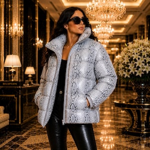 May include: A puffer jacket featuring a grey and black snakeskin print, a high collar, and a front zipper closure. The jacket has long sleeves with elastic cuffs and two side pockets. It is worn over a black top and black textured pants.