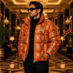 Könnte beinhalten: Eine leuchtend orangefarbene Daunenjacke mit einem detaillierten schwarzen Schlangenmuster. Die Jacke hat einen hohen Kragen, einen durchgehenden Frontreißverschluss und lange Ärmel. Sie wird über einem schwarzen Rollkragenpullover getragen.