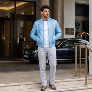 May include: A light blue bomber jacket with a textured snakeskin pattern, worn over a plain white t-shirt and light gray trousers. The outfit is completed with brown leather loafers, creating a coordinated look for a professional setting.