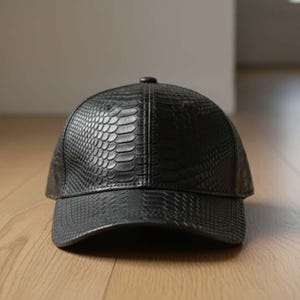 May include: A black baseball cap with a textured, snakeskin-like pattern. The cap has a curved brim and a small button on top. The hat is made of a leather-like material and sits on a wooden floor.