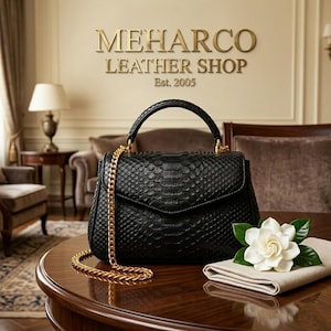 May include: A black handbag with a textured snakeskin pattern, a top handle, and a gold chain shoulder strap. The bag features a flap closure. Next to the bag sits a folded beige cloth topped with a white gardenia flower with green leaves.