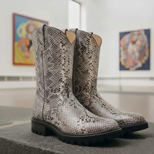 Może przedstawiać: Para wysokich kowbojskich butów ze wzorem skóry węża w odcieniach szarości i bieli. Buty mają zaokrąglony nosek, obcas i czarną gumową podeszwę. Cholewka butów ma około 30 cm wysokości.