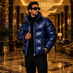 May include: A glossy, royal blue puffer jacket featuring a textured snakeskin pattern, a high collar, and a front zip fastening. The jacket is worn over a black turtleneck jumper and black trousers, paired with dark sunglasses and a gold wristwatch.