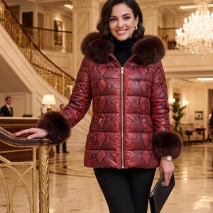 Könnte beinhalten: Eine rote und schwarze Steppjacke mit Schlangenledermuster, ausgestattet mit einer großen, braun fellbesetzten Kapuze und passenden Fellbündchen an den Ärmeln. Die Jacke hat einen goldenen Reißverschluss und zwei seitliche Reißverschlusstaschen, getragen zu einer schwarzen Hose.