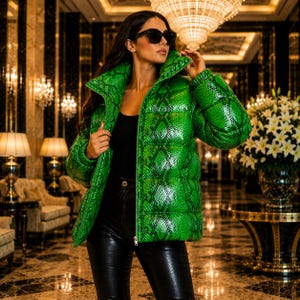 May include: A bright green puffer jacket featuring a black snakeskin pattern, a high collar, and a front zip closure. The jacket is worn over a black top and black trousers, paired with black sunglasses, showcasing a glossy, textured finish.