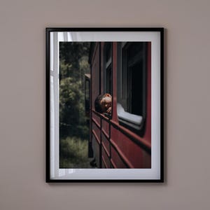 Può includere: Stampa incorniciata con una persona che guarda fuori da un finestrino di un treno rosso. L'esterno del treno è rosso scuro, con finestre e ringhiere visibili. Lo sfondo rivela alberi verdi, suggerendo un viaggio in treno pittoresco.