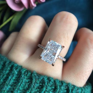 May include: A rose gold ring featuring a large, rectangular radiant-cut clear gemstone held by four prongs. The thin band is encrusted with small, sparkling clear stones along the visible portion of the shank, creating a bright, reflective appearance.
