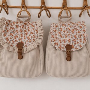 Könnte beinhalten: Zwei beigefarbene Rucksäcke aus Cord mit einer Blumenmuster-Klappe in Orange und Weiß. Der linke Rucksack hat einen Rüschenbesatz an der Klappe. Beide Taschen verfügen über einen braunen Kunstlederriemen mit goldener Schnalle und einen Tragegriff.