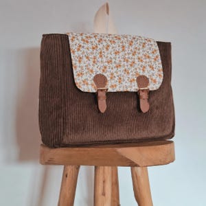 Peut inclure: Un sac à dos en velours côtelé marron doté d'un rabat blanc à motif floral jaune et orange. Le sac comprend une poignée supérieure et deux lanières en similicuir marron avec des boucles métalliques pour fermer le rabat avant.