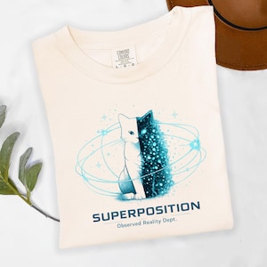 May include: Cream-colored t-shirt featuring a graphic of a cat split in half, one side white, the other a dark blue with a starry effect. The shirt has the word "SUPERPOSITION" and "Observed Reality Dept." printed below the cat design.