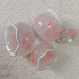 May include: A set of decorative glass items, including small bowls and a vase, featuring a pink floral pattern. The glass is translucent, with white rims. The items are displayed on a white, textured surface.