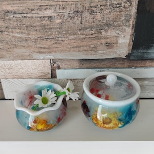 May include: Two decorative ceramic containers with floral designs. One is open, holding white daisies. The other is a lidded jar. Both have a white rim and a colourful, abstract floral pattern in shades of blue, yellow, and red.