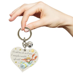Può includere: Portachiavi a forma di cuore con un'illustrazione a cartoni animati di un ragazzo su un aereo, una volpe e testo in italiano. Il portachiavi ha un anello argentato e un piccolo ciondolo a campana. Il testo recita "Si vede bene solo con il cuore, l'essenziale è invisibile agli occhi!"