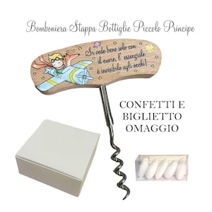 Può includere: Un cavatappi in legno con una spirale in metallo, con un'illustrazione dipinta di un personaggio e un aereo. Il testo "Si vede bene solo con il cuore, l'essenziale è invisibile agli occhi!" è sull'impugnatura in legno. Include una scatola bianca e caramelle.