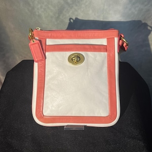 May include: A rectangular crossbody bag in white, coral, and gold. The bag features a white center panel, coral-colored trim, and a coral-colored strap. A gold-colored clasp secures the front, and a coral-colored tag hangs from the strap.