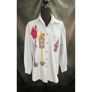 May include: Light blue denim shirt with long sleeves and a button-down front. The shirt features colorful, appliqued birdhouse designs in red, yellow, and blue. A small pocket is on the left chest. The collar has a white floral pattern.