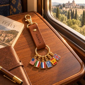 May include: A brown leather keychain with a gold-tone metal clasp and ring. The leather strap is embossed with the word ROAMER. Attached to the gold ring are six small rectangular enamel charms featuring the flags of the USA, Italy, France, Greece, Spain, and the UK.