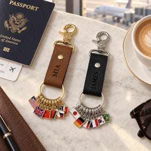 May include: Two leather keychains with metal lobster clasps and rings. One brown leather strap is embossed with the name MIA and features gold-toned flag charms. One black leather strap is embossed with the name JAMES and features silver-toned flag charms.