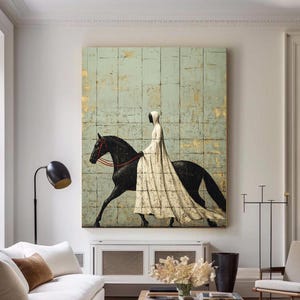 May include: An abstract painting depicts a figure in a white gown riding a black horse. The artwork is set against a textured, light blue and gold background. The painting hangs above a white cabinet in a living room.