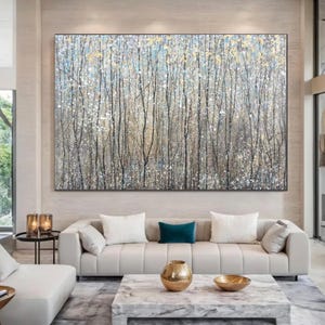 May include: Large abstract painting of a forest scene with tall, slender trees in grey, black, and gold. The artwork has a textured appearance and is mounted above a cream sofa in a modern living room setting.