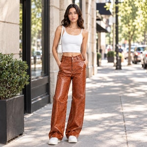 May include: A white sleeveless crop top paired with high-waisted, wide-leg trousers in a glossy, brown, crocodile-embossed texture. The trousers feature a button and zip fly closure, side pockets, and a full-length cut that reaches the ground.