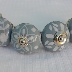 May include: A collection of six grey ceramic cabinet knobs, each adorned with a white floral design. The knobs feature silver-toned metal bases and decorative centres. The knobs are arranged in a semi-circle.