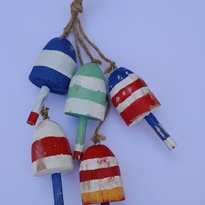 May include: Five small, painted wooden buoys in shades of blue, red, and green hang from a natural twine rope. Each buoy features a different design, such as stripes or solid colours, and has a distressed, rustic appearance, giving a nautical theme.