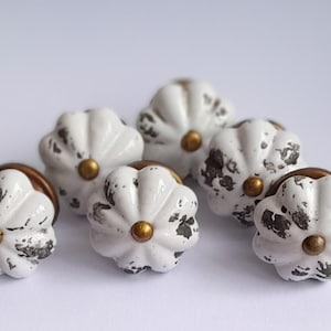 May include: Six white ceramic cabinet knobs with a distressed finish and gold-coloured accents. The knobs have a flower-like shape and are arranged on a white surface. These decorative knobs are ideal for adding a vintage touch to furniture.