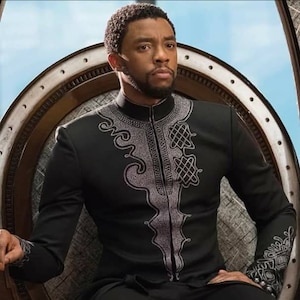 May include: A man wearing a black long-sleeved shirt with silver embroidery. The shirt has a high collar and a zip down the front. The man is sitting on a throne.