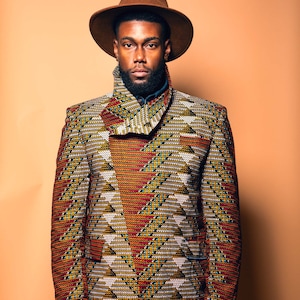 K&ouml;nnte beinhalten: Ein Mann tr&auml;gt einen braunen Fedora und eine gemusterte Jacke mit einem geometrischen Design in Rot-, Gelb- und Wei&szlig;t&ouml;nen. Die Jacke hat einen hohen Kragen und einen Knopfverschluss.