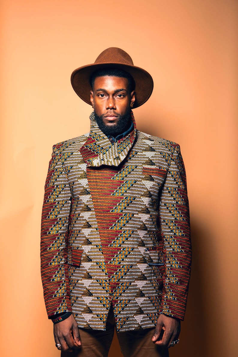 K&ouml;nnte beinhalten: Ein Mann tr&auml;gt einen braunen Fedora und eine gemusterte Jacke mit einem geometrischen Design in Rot-, Gelb- und Wei&szlig;t&ouml;nen. Die Jacke hat einen hohen Kragen und einen Knopfverschluss.
