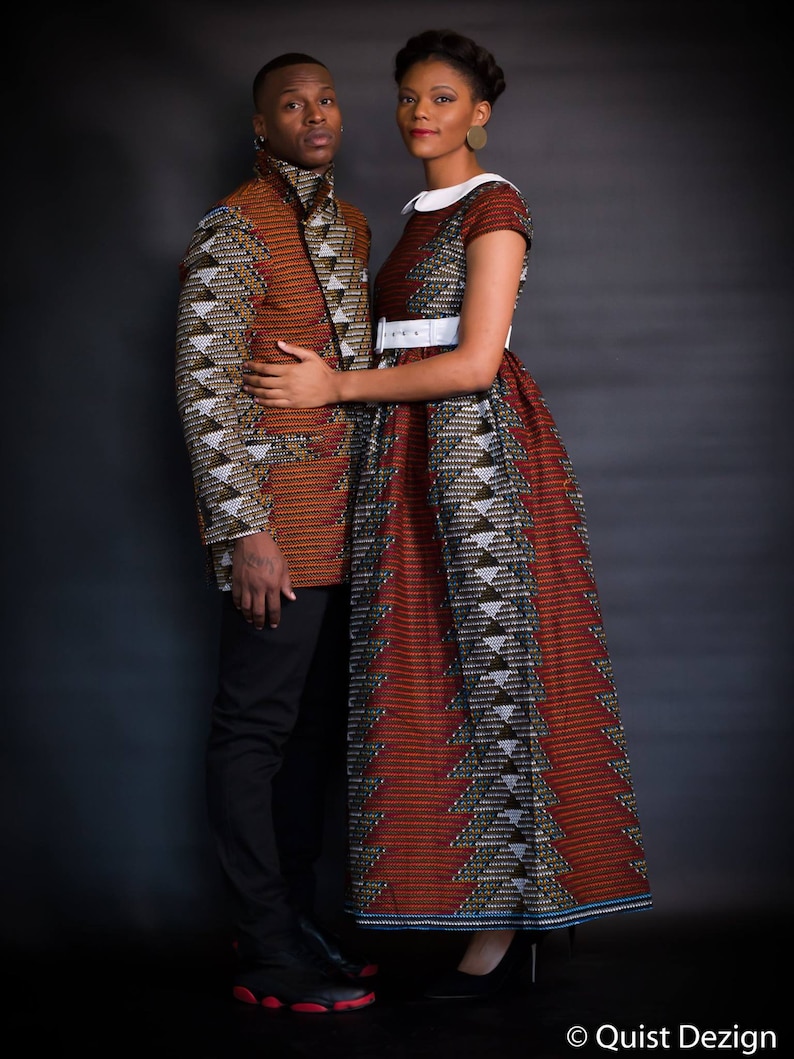 K&ouml;nnte beinhalten: Ein Paar tr&auml;gt passende gemusterte Outfits. Die Frau tr&auml;gt ein rot-schwarzes Kleid mit einem wei&szlig;en G&uuml;rtel und einem wei&szlig;en Kragen. Der Mann tr&auml;gt eine braun-schwarze Jacke mit einem &auml;hnlichen Muster. Sie stehen vor einem dunklen Hintergrund.