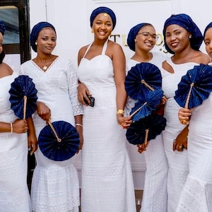 May include: Six women wearing white lace dresses and matching blue head wraps hold blue and gold hand fans. They are standing in front of a white wall.