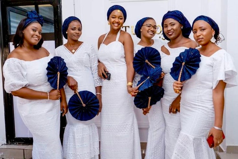 May include: Six women wearing white lace dresses and matching blue head wraps hold blue and gold hand fans. They are standing in front of a white wall.