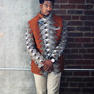 K&ouml;nnte beinhalten: Ein Mann tr&auml;gt einen gemusterten orangefarbenen, wei&szlig;en und schwarzen Blazer mit geometrischem Muster. Er tr&auml;gt eine beige Hose und eine Uhr am linken Handgelenk.