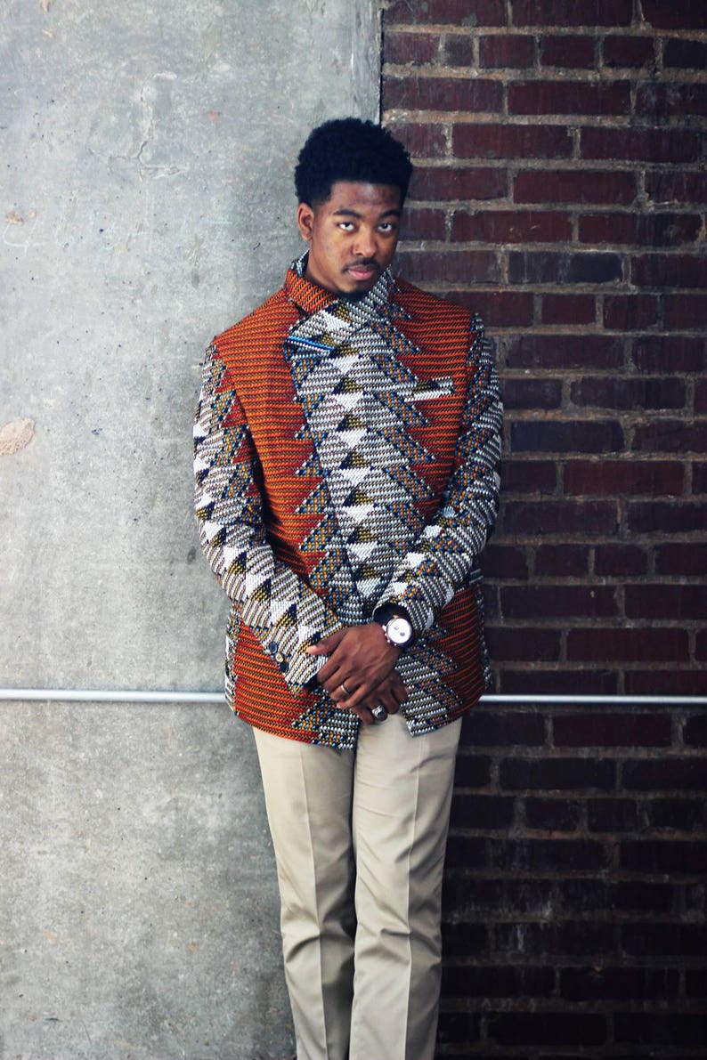 K&ouml;nnte beinhalten: Ein Mann tr&auml;gt einen gemusterten orangefarbenen, wei&szlig;en und schwarzen Blazer mit geometrischem Muster. Er tr&auml;gt eine beige Hose und eine Uhr am linken Handgelenk.