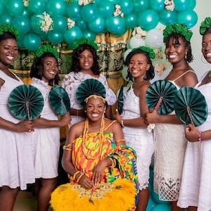 May include: A group of seven women in white dresses and green fans, with one woman in the center wearing a colorful kente cloth dress and a gold headpiece. They are standing in front of a wall decorated with green and white balloons.