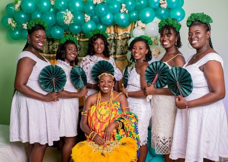 May include: A group of seven women in white dresses and green fans, with one woman in the center wearing a colorful kente cloth dress and a gold headpiece. They are standing in front of a wall decorated with green and white balloons.