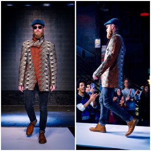 K&ouml;nnte beinhalten: Ein Mann tr&auml;gt eine gemusterte Jacke, blaue Jeans, braune Schuhe und einen blauen Hut. Er geht auf einem Laufsteg entlang, w&auml;hrend eine Menschenmenge zuschaut.
