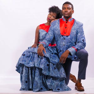 Puede incluir: Una pareja vistiendo atuendos a juego de color azul y rojo con estampado. La mujer lleva un vestido con falda de volantes y el hombre lleva una chaqueta con cuello rojo. Ambos miran a la c&aacute;mara.