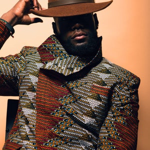 K&ouml;nnte beinhalten: Ein Mann tr&auml;gt einen braunen Fedora-Hut und eine gemusterte Jacke mit hohem Kragen. Die Jacke hat ein geometrisches Muster in Rot-, Orange-, Gelb-, Blau- und Wei&szlig;t&ouml;nen.