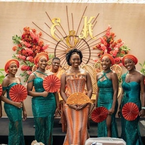 May include: A group of women in teal dresses and orange head wraps stand in front of a floral backdrop. They are holding orange fans and smiling. The bride is in the center wearing a patterned orange dress.