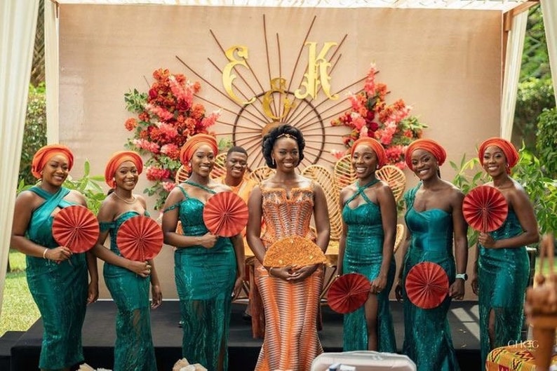 May include: A group of women in teal dresses and orange head wraps stand in front of a floral backdrop. They are holding orange fans and smiling. The bride is in the center wearing a patterned orange dress.