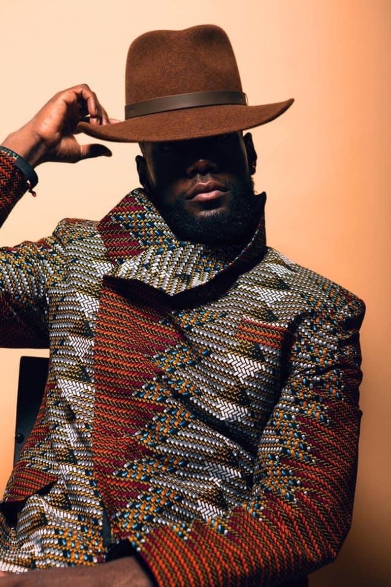 K&ouml;nnte beinhalten: Ein Mann tr&auml;gt einen braunen Fedora-Hut mit einem braunen Lederband und eine gemusterte Jacke in den Farben Rot, Orange, Gelb, Wei&szlig; und Blau.