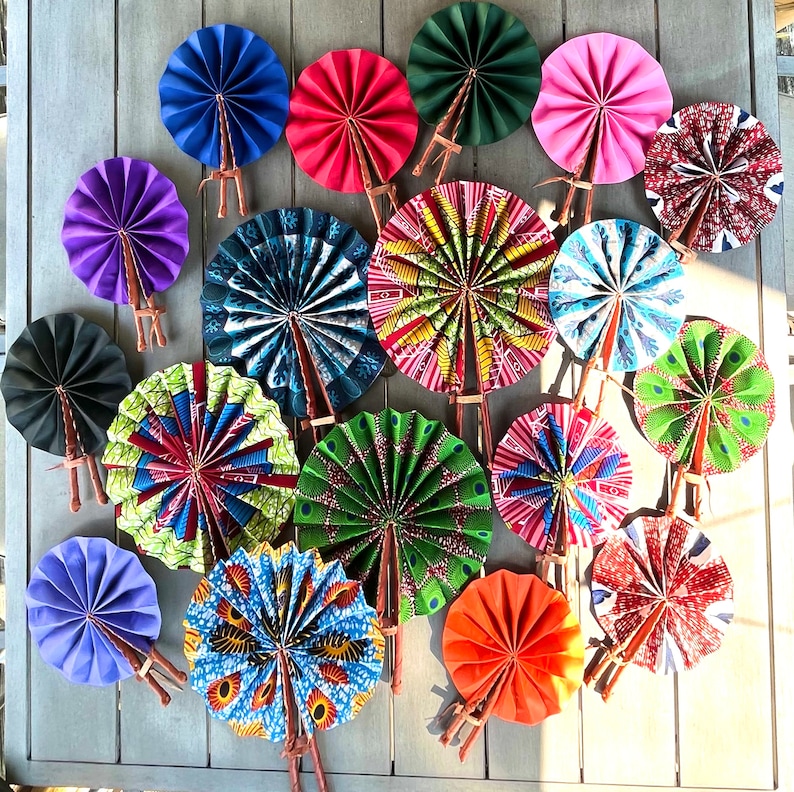 May include: A collection of colorful hand fans with various patterns and designs. The fans are made of paper and have wooden handles. The fans are arranged in a circle on a wooden surface.