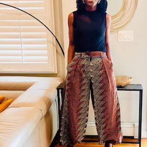 K&ouml;nnte beinhalten: Eine Frau tr&auml;gt ein schwarzes &auml;rmelloses Oberteil mit schwarzem Mesh-&Uuml;berzug, eine breite Hose mit braunen und goldenen Mustern und eine schwarze Sonnenbrille. Sie steht in einem Raum mit einem wei&szlig;en Sofa und einem Holzboden.