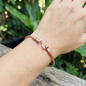 May include: A copper-colored metal cuff bracelet featuring a central S-shaped wire detail flanked by two sections of tightly coiled wire wrapping. The bracelet has a smooth, rounded band design that maintains a consistent metallic finish throughout.